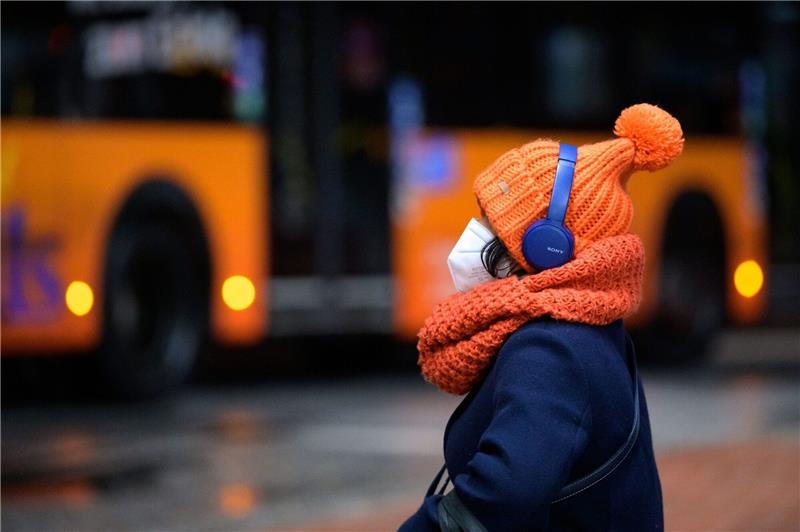 Streit über Vorstoß zum Ende der Maskenpflicht im Nahverkehr Eine Frau sitzt mit FFP2-Maske, dicker Mütze und Kopfhörern an einem Busbahnhof, während ein Linienbus vorbeifährt.
