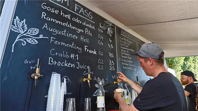Mann mit grauer Kappe zapft Bier an einer schwarzen Tafel mit handgeschriebener Getränkekarte und Zapfhähnen.
