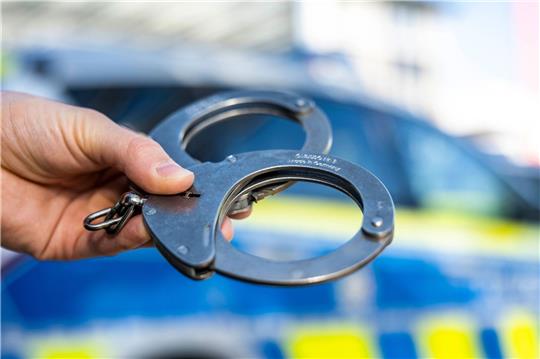 Eine Hand hält Handschellen vor einen Streifenwagen der Polizei.