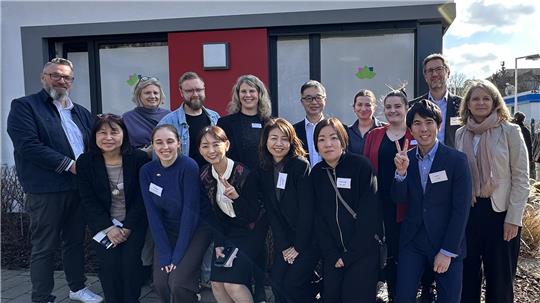 Gruppe von Menschen mit Namensschildern steht vor einem modernen Gebäude mit roter Tür und Fenstern bei sonnigem Wetter.