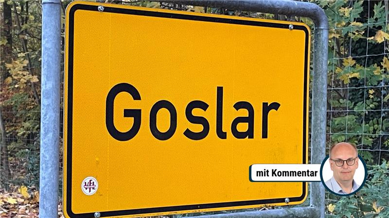 Gelbes Ortsschild mit schwarzer Umrandung und der schwarzen Aufschrift 'Goslar' vor einem Waldzaun und Bäumen