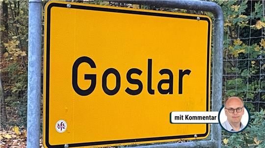 Gelbes Ortsschild mit schwarzer Umrandung und der schwarzen Aufschrift 'Goslar' vor einem Waldzaun und Bäumen