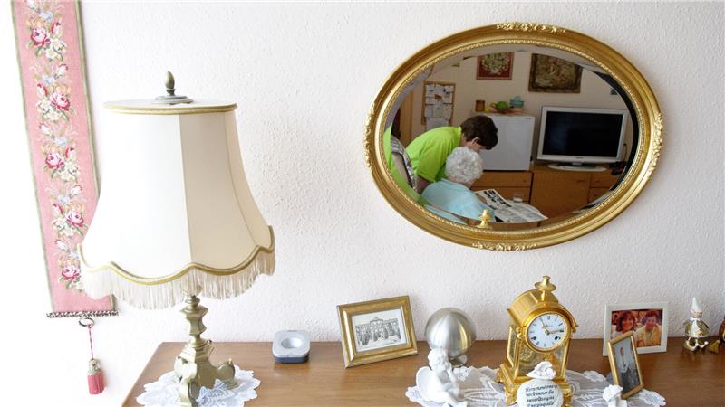 In einem Spiegel ist zu sehen, wie eine Pflegerin sich um eine Seniorin kümmert.