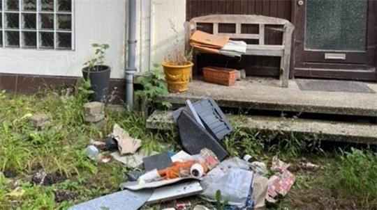 Vor einer Haustür liegt verstreut Müll mit einem umgekippten Mülleimer und mehreren Blumentöpfen auf einer kleinen Treppe und im Gras