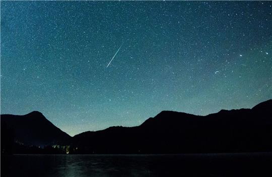 Eine Sternschnuppe leuchtet neben der Milchstraße am Himmel über dem Walchensee.