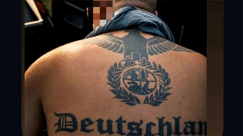 Rückenansicht eines Mannes mit großem schwarzen Tattoo eines Adlers über einem Lorbeerkranz mit einem runden Symbol in der Mitte.
