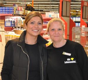 Zwei Frauen stehen in einem großen, gut beleuchteten Supermarkt mit roten Regalen und Einkaufswagen im Hintergrund.