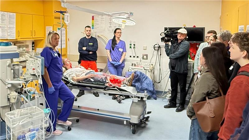 Blick in ein Zimmer im Krankenhaus: Auf einem Bett liegt eine Patientin, drumherum stehen Krankenhausmitarbeiter und Schüler.