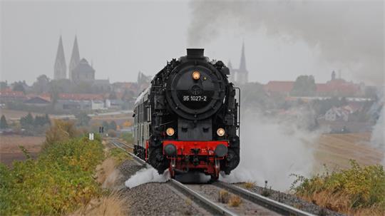Eine Dampflok fährt auf den Schienen. Im Hintergrund ist Magdeburg mit seinen Kirchen zu sehen.