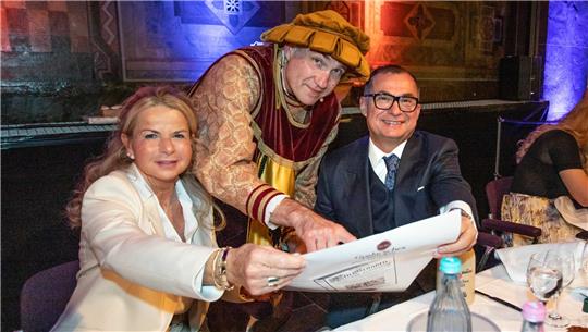 Drei Personen sitzen an einem gedeckten Tisch, eine Person in historischer Kleidung zeigt auf ein großes Blatt Papier, die anderen beiden schauen darauf