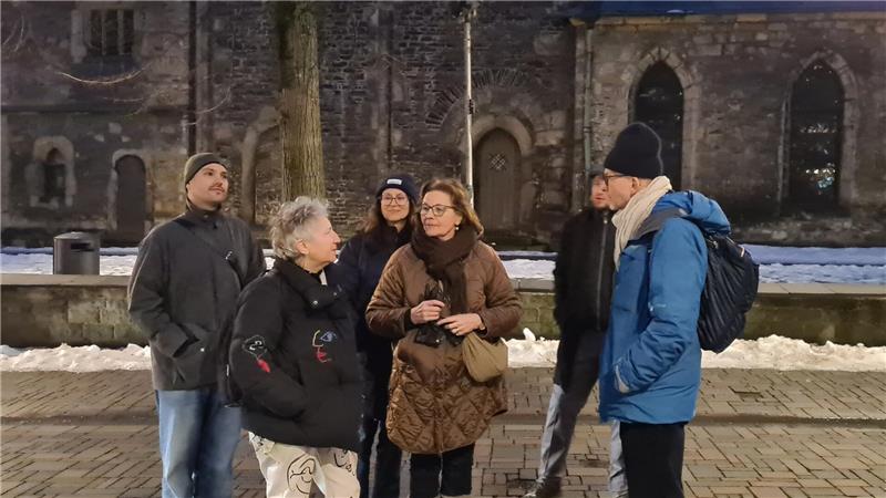 Gruppe von sechs Personen in Winterkleidung steht auf gepflastertem Platz vor einer alten Kirche mit gotischen Fenstern.