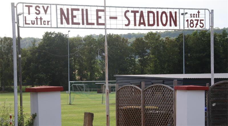 Eingangstor mit Schild 'TSV Lutter NEILE - STADION seit 1875' vor einem Fußballfeld mit Toren und Bäumen im Hintergrund. 
