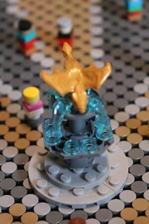 Goldfarbener Vogel auf einem Sockel aus transparenten blauen und grauen Bausteinen auf einer grauen Grundplatte mit Noppen.