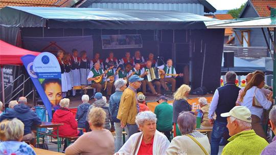 Eine Brauchtumsgruppe musiziert auf einer Bühne vor Menschen.