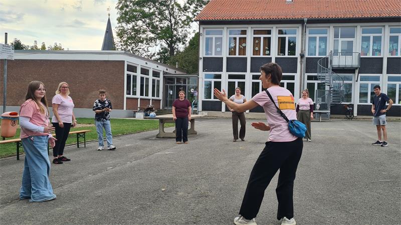 Einige Personen stehen im Keis auf einem Schulhof und blicken auf eine Trainerin, die Bewegungen vormacht. Im Hintergrund die Kirchturmspitze.