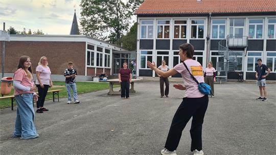 Einige Personen stehen im Keis auf einem Schulhof und blicken auf eine Trainerin, die Bewegungen vormacht. Im Hintergrund die Kirchturmspitze.