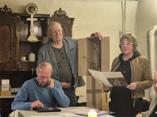 Drei Menschen vor einem alten Schrank. Eine Frau guckt in ein Faltblatt, ein Mann legt am Tisch die Brille zurecht, im Hintergrund hält jemand einen Pappkarton.