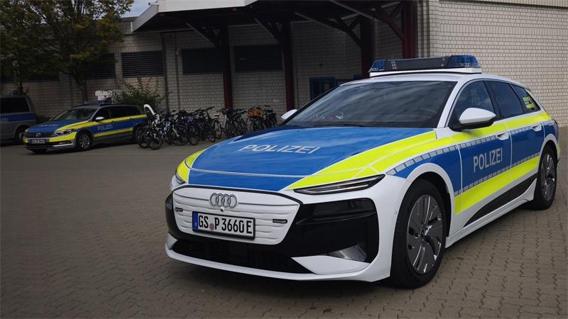 Ein weiß-blau-gelb beklebtes Polizeifahrzeug steht auf dem Hof eines Gebäudes. Im Hintergrund sind zwei weitere Polizeifahrzeuge, Fahrräder und eine Fahrzeughalle zu sehen.