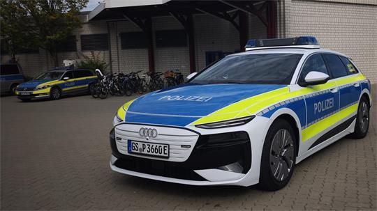 Ein weiß-blau-gelb beklebtes Polizeifahrzeug steht auf dem Hof eines Gebäudes. Im Hintergrund sind zwei weitere Polizeifahrzeuge, Fahrräder und eine Fahrzeughalle zu sehen.