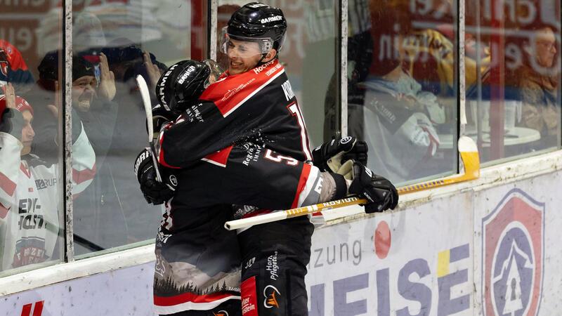 Zwei Eishockeyspieler in schwarzer Ausrüstung umarmen sich jubelnd an der Bande vor Zuschauern hinter der Scheibe