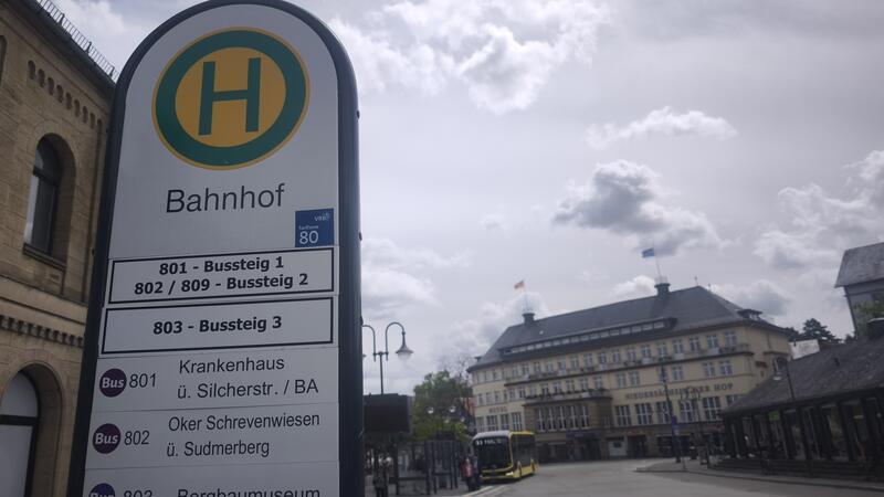 Haltestellenschild mit dem Schriftzug 'Bahnhof' und Buslinieninformationen vor einem historischen Bahnhofsgebäude und einer Straße mit wartendem Bus