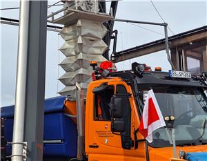 Oranger Lastwagen mit blauem Anhänger, an der Fahrerseite eine weiße Flagge mit rotem Kreuz, im Hintergrund ein hoher Mast mit einer weißen, faltbaren Struktur.