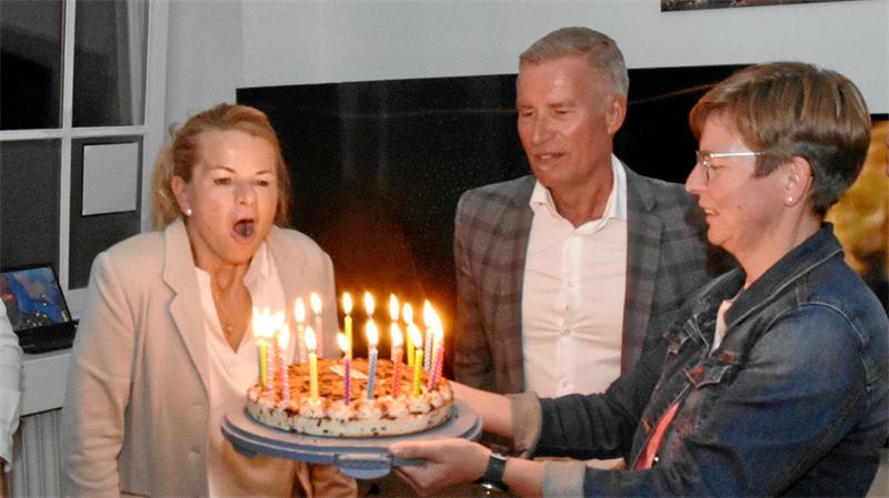 Drei Personen, eine Frau bläst Kerzen auf einer Geburtstagstorte aus, eine andere Frau hält die Torte, ein Mann steht daneben in einem Raum mit Fenster und Kamin.