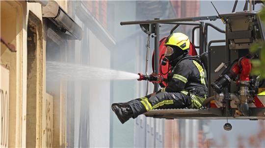 Ein Feuerwehrmann in einer Drehleiter hält einen Schlauch in ein geöffnetes oder zerstörtes Fenster.