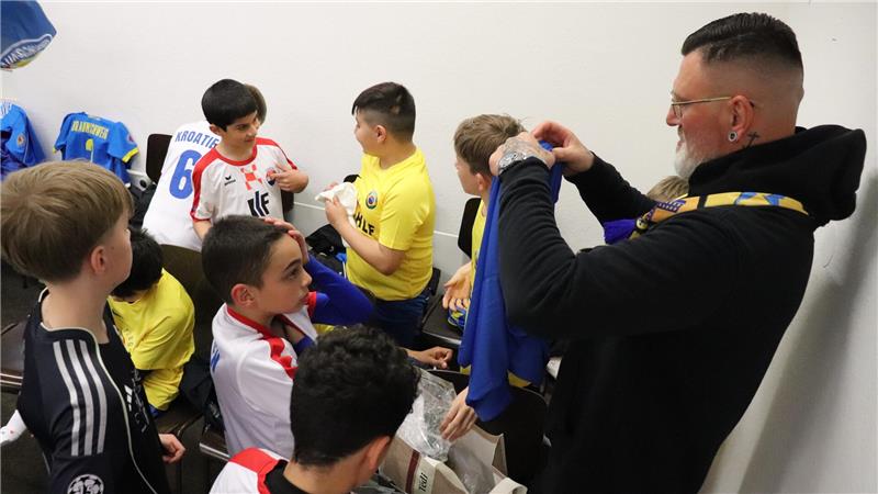 Gruppe von Jungen in Sportkleidung in einem Umkleideraum, ein Erwachsener hält ein blaues Trikot hoch