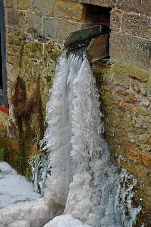 Wasserfall, der aus einem rechteckigen Auslass an einer alten Steinmauer fließt und teilweise zu Eis gefroren ist