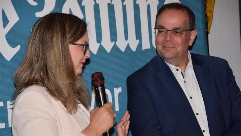 Zwei Personen im Gespräch, eine Frau mit Brille hält ein Mikrofon, ein Mann mit Brille schaut sie an, beide vor einem blauen Hintergrund mit weißer Schrift.