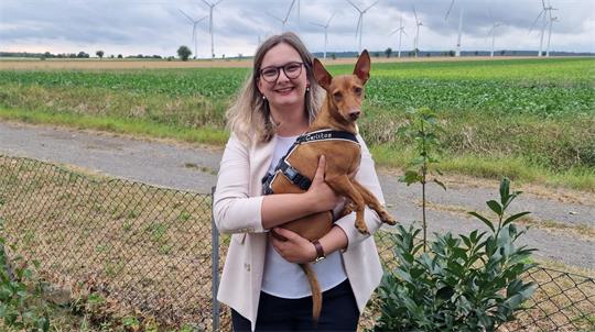 Frau mit Brille und hellem Blazer hält kleinen Hund mit aufgerichteten Ohren vor einem Feld mit Windrädern.