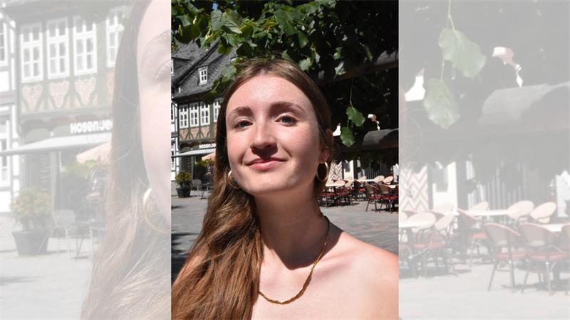 Junge Frau mit langen braunen Haaren und weißen Trägeroberteil sitzt vor einer Kirche mit Sandsteinmauer und gotischen Fenstern.