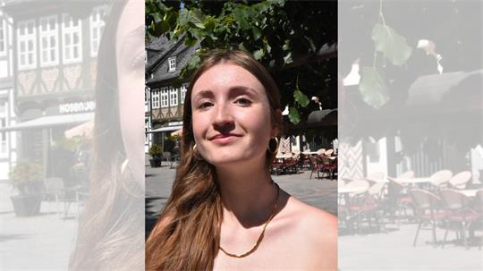 Junge Frau mit langen braunen Haaren und weißen Trägeroberteil sitzt vor einer Kirche mit Sandsteinmauer und gotischen Fenstern.