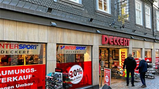 Fassade eines Sportgeschäfts mit roten Werbeschildern für einen 20-Prozent-Rabatt und zwei Personen vor dem Eingang auf gepflastertem Gehweg. 