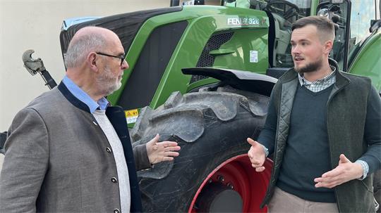 Zwei Männer stehen vor einem großen grünen Traktor und gestikulieren im Gespräch