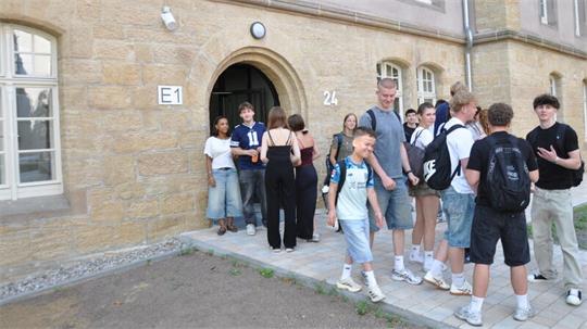 Gruppe Jugendlicher mit Rucksäcken steht und geht vor einem Sandsteingebäude mit Bogenportal und der Hausnummer 24