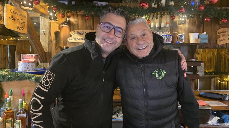 Zwei Männer in Winterjacken stehen in einer festlich dekorierten Holzhütte mit Weihnachtskugeln und Lichterketten, im Hintergrund Regale mit Flaschen und handgeschriebene Preisschilder.