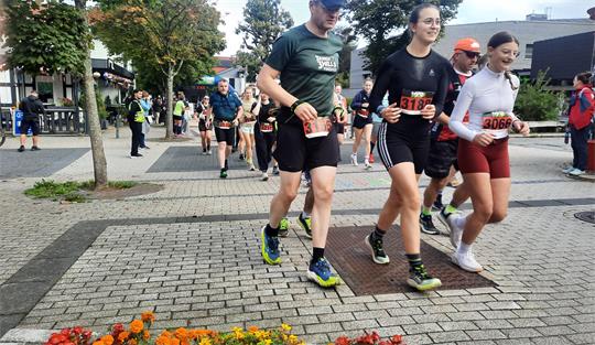 Gruppe von Läufern mit Startnummern läuft auf gepflastertem Weg an einem Blumenbeet vorbei in einer städtischen Umgebung mit Bäumen und Gebäuden im Hintergrund