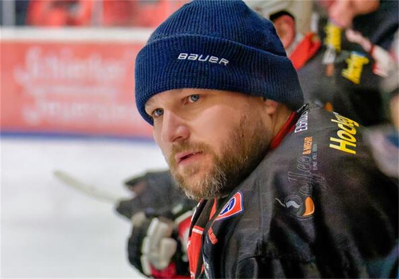 Eishockeyspieler mit blauer Mütze und schwarzem Trikot steht an der Bande und blickt nach vorne