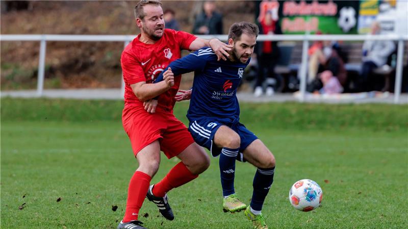 Zwei Fußballspieler in Aktion, einer in rotem Trikot, der andere in blauem Trikot, kämpfen um den Ball auf dem Spielfeld
