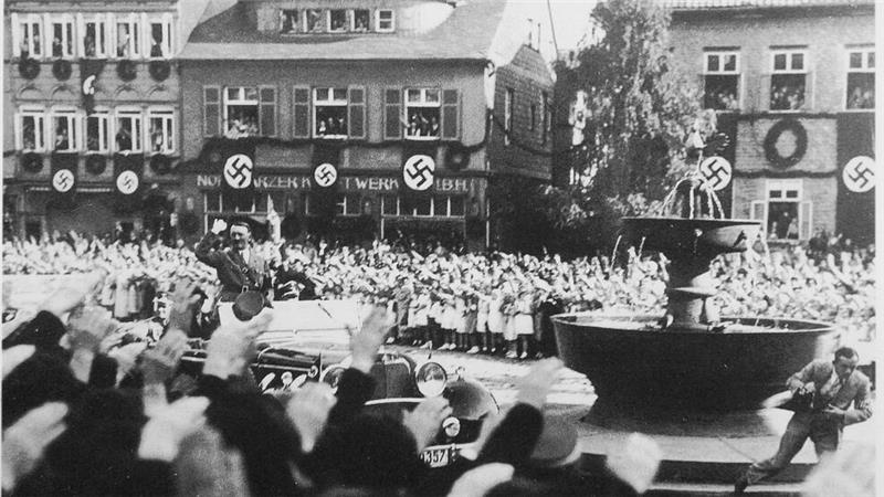 Große Menschenmenge auf dem Goslarer Marktplatz mit Hakenkreuzfahnen an Gebäuden. Viele Personen heben die rechte Hand zum Gruß, ein Auto mit Adolf Hitler fährt durch die Menge.