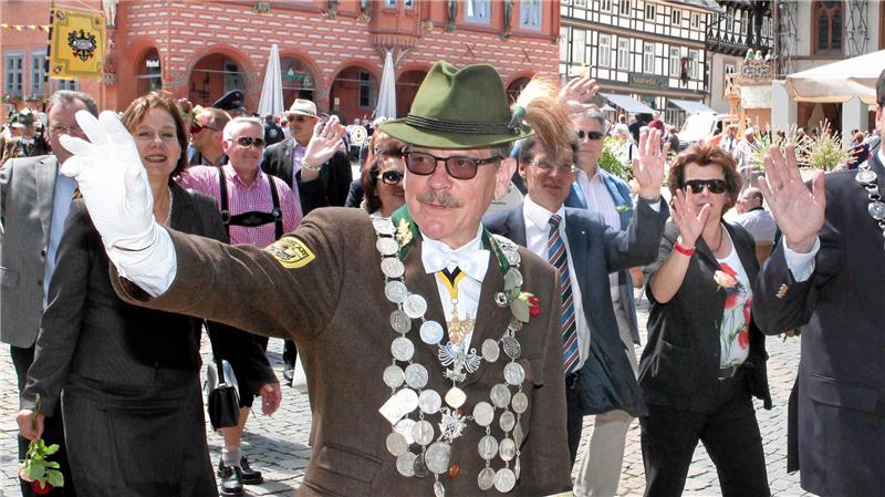 Person in traditioneller Tracht mit grünem Hut und großer silberner Kette hebt die rechte Hand vor einer Gruppe von Menschen auf einem historischen Marktplatz
