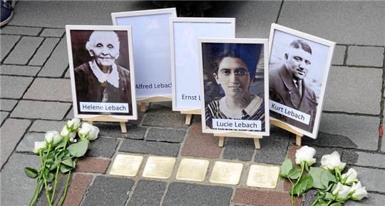 Gedenkstelle mit Porträtfotos auf Staffeleien und mehreren weißen Rosen auf dem Boden vor fünf Stolpersteinen auf Pflastersteinen