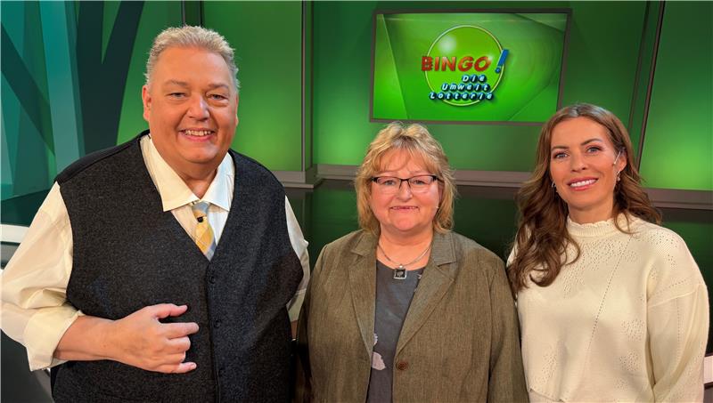 Drei Personen lachen, im Hintergrund zeigt ein Bildschirm "Bingo! Die Umweltlotterie" an.