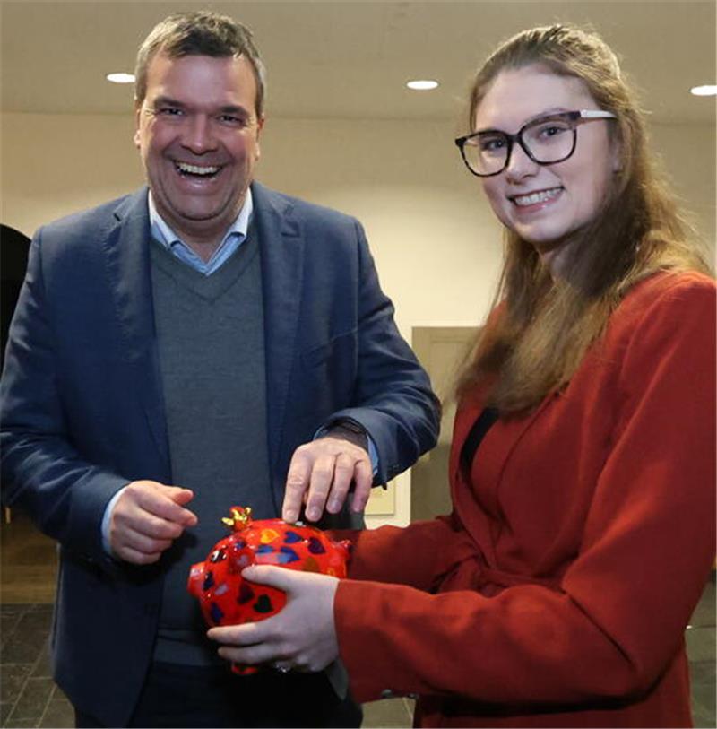 Zwei Personen stehen in einem Innenraum, eine hält eine rote Spardose in Form eines Sparschweins mit gelben Punkten.