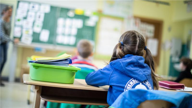Schülerin mit Zöpfen und blauem Kapuzenpullover sitzt an einem Schultisch mit grünem Mäppchen und blickt auf eine Lehrerin vor einer Tafel im Klassenraum