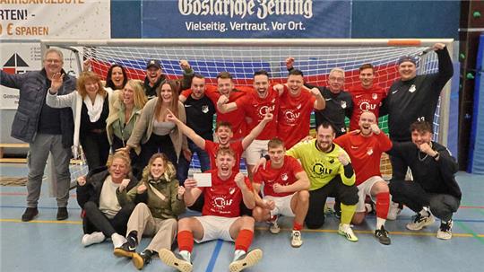Gruppenfoto einer Hallenfußballmannschaft mit Spielern in roten und schwarzen Trikots vor einem Tor, einige sitzen, andere stehen und jubeln in einer Sporthalle