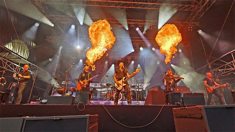 Band spielt auf Bühne mit Pyrotechnik und zwei großen Feuersäulen über den Musikern. 