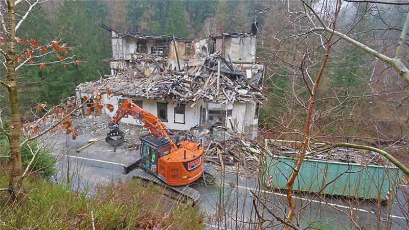 Bagger reißt teilweise eingestürztes Haus in bewaldeter Umgebung ab.